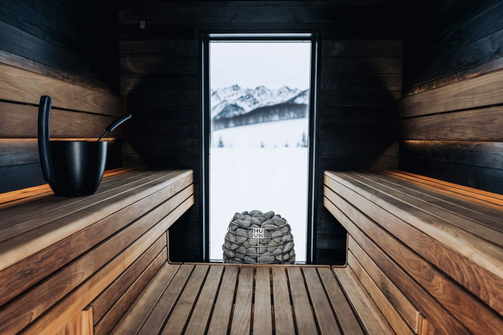Dark wood sauna interior with a heater full of stones and a view over snow-capped mountains
